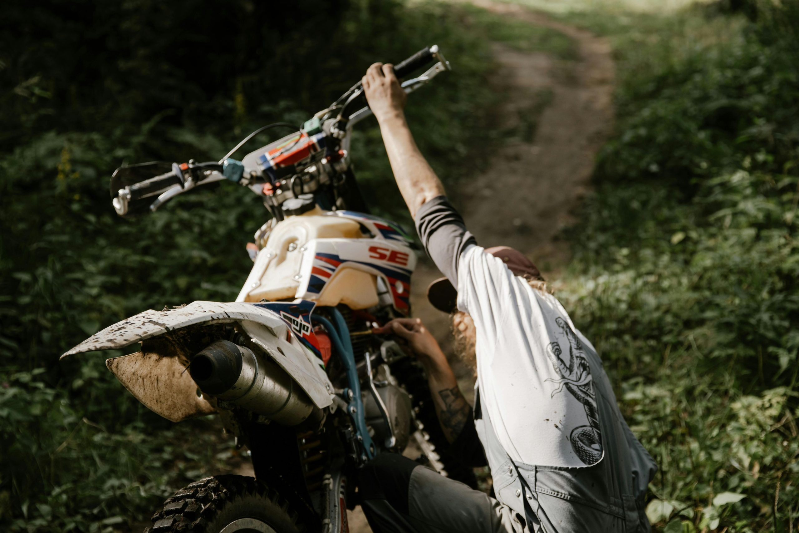 A bearded mechanic repairs a motocross bike on a forest path during the day.